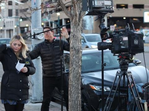 A television reporter prepares for a live report beside a camera crew on a city street.