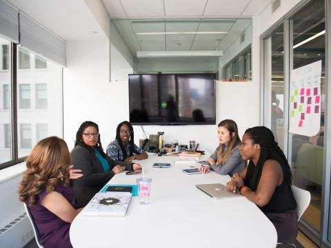 Five women collaborate around a table in a modern office meeting.