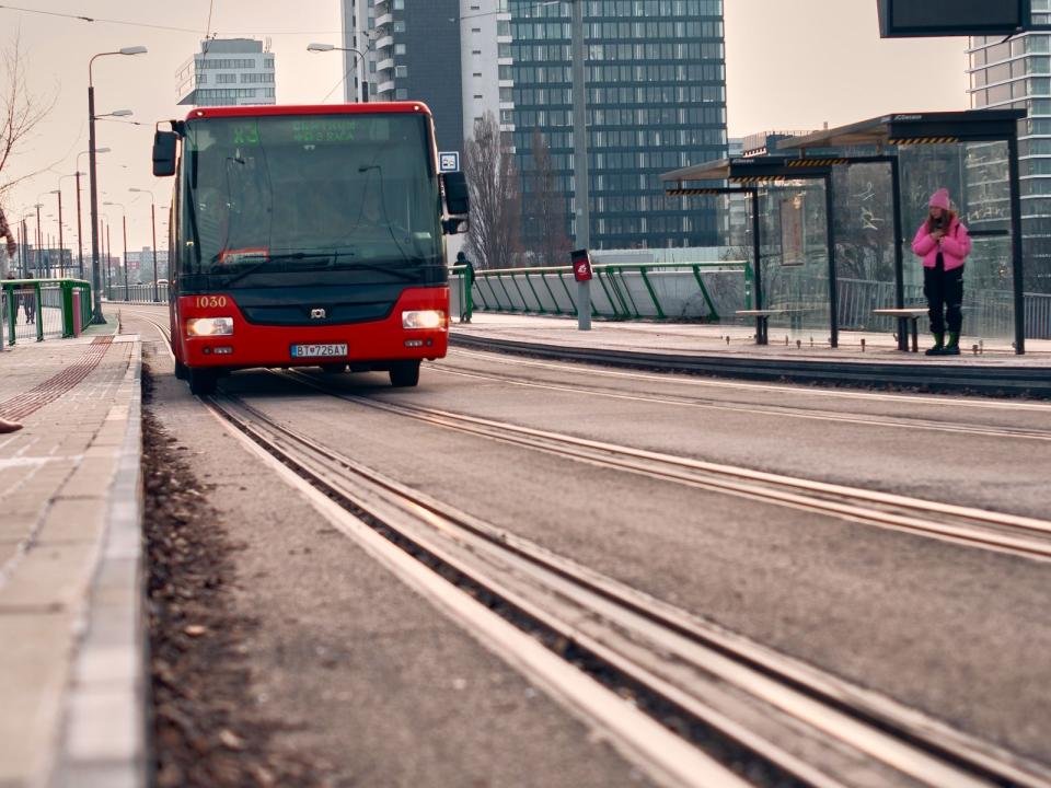 A red city bus approaches a tram stop between modern buildings.