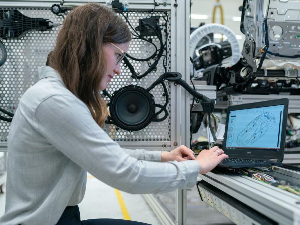 A woman works at a laptop beside electronic equipment in an engineering workshop.
