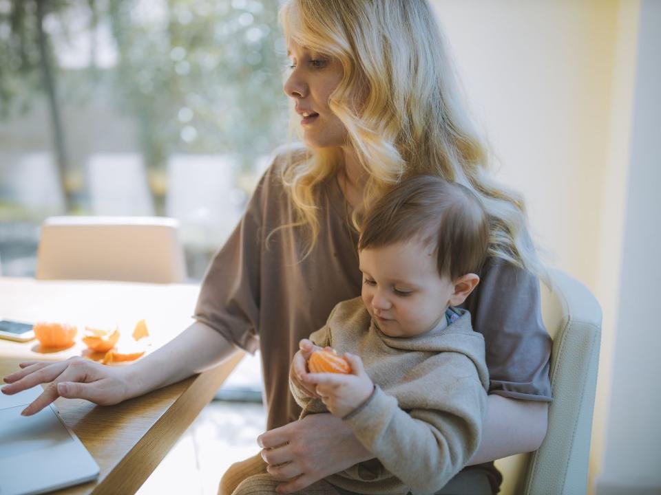 A mother works on a laptop while holding her young child at home.