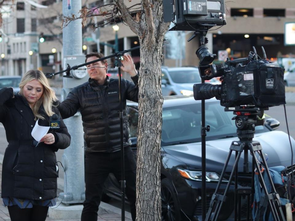 A television reporter prepares for a live report beside a camera crew on a city street.