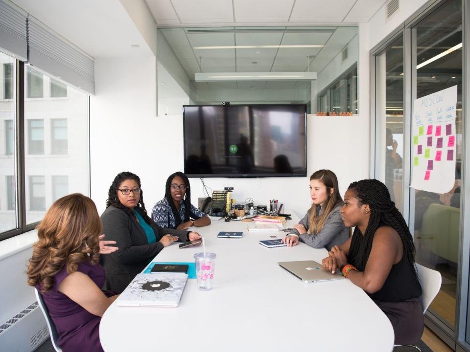 Five women collaborate around a table in a modern office meeting.